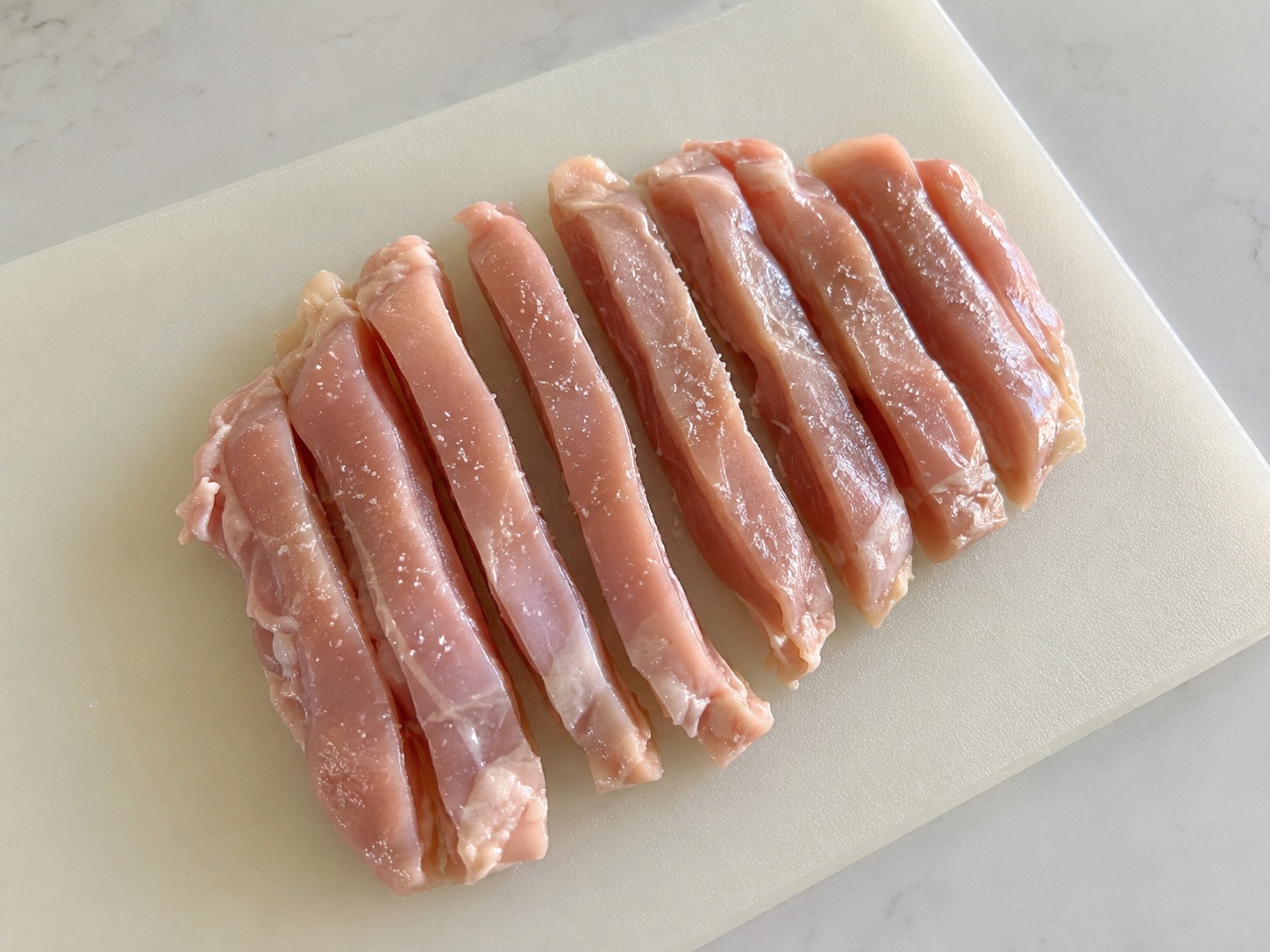 Boneless chicken thighs cut lengthwise into lightly salted strips on a cutting board